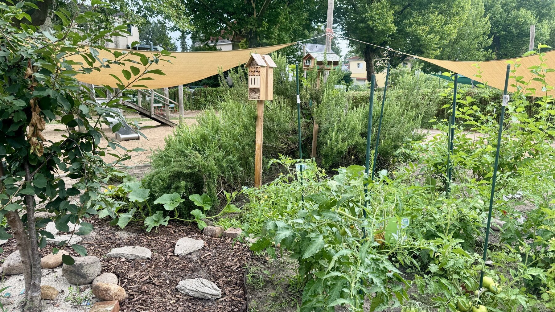Zu sehen sind Beete mit Insektenhotel, Vogelhaus und Sonnensegeln im Hintergrund im Garten der Humanistischen Kita Friedrich-Engels-Straße in Berlin-Pankow.