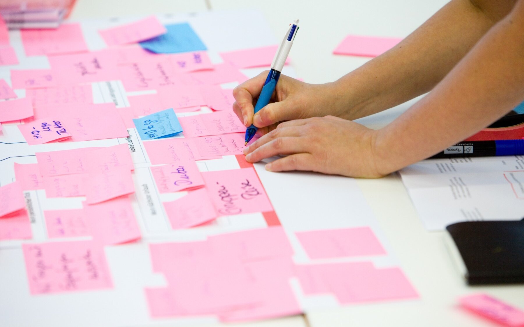 Auf einem Tisch liegen weiße Papierblätter, auf denen beschriebene rosa und blaue Post-its kleben. Eine Person, von der nur die Unterarme zu sehen sind, schreibt mit einem Kugelschreiber etwas auf ein rosa Post-it.
