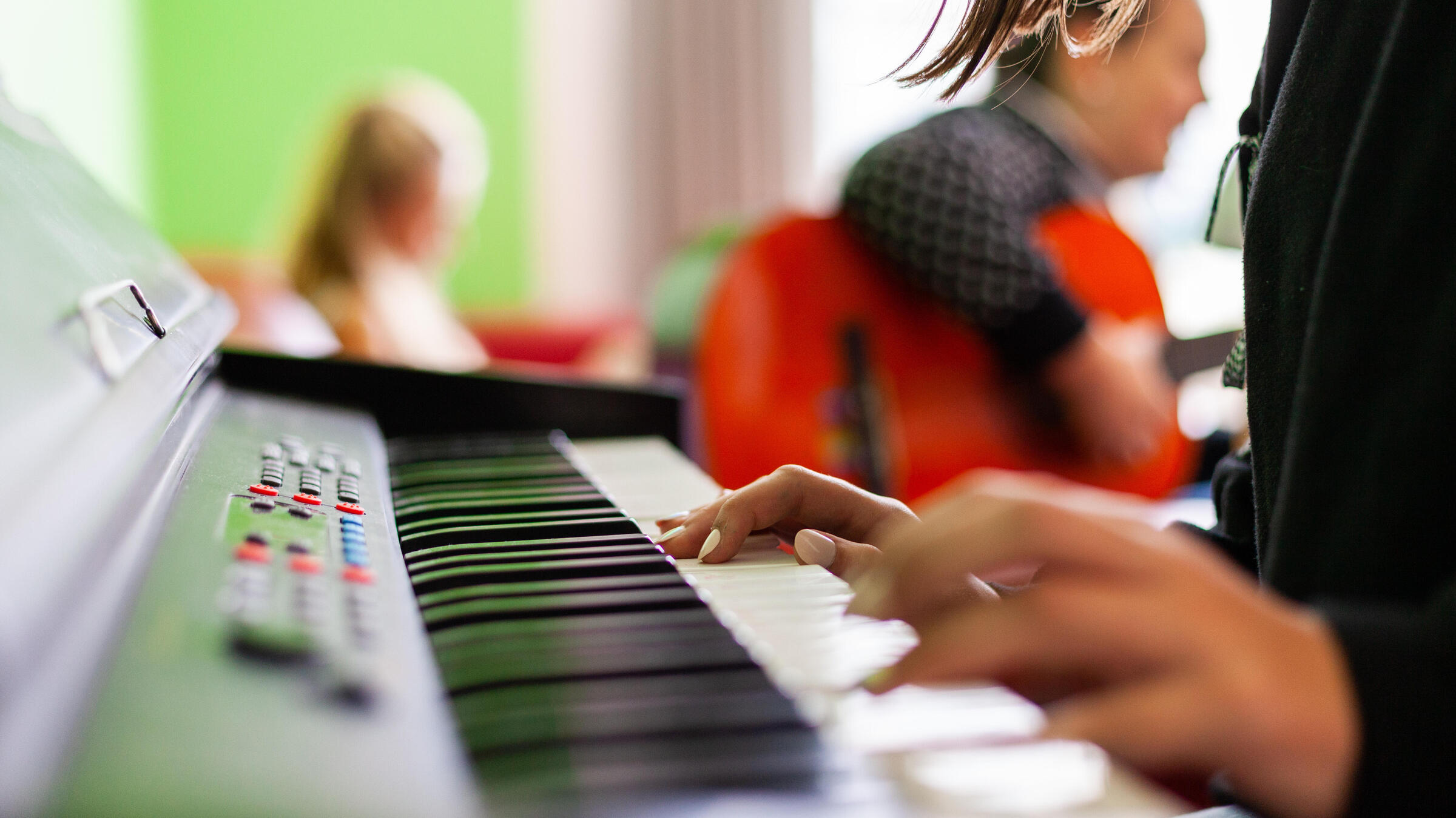 Zu sehen sind Hände, die auf einem Keyboard spielen. Im Hintergrund ist eine verschwommene Person zu erkennen, die auf einer Gitarre spielt.