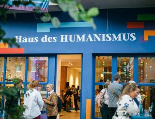 Haus des HUMANISMUS Potsdamer Straße WHT 2022 Menschen stehen in feierliche Stimmung vor und in einem Veranstaltungsort mit blauer Fassade und Schriftzug: Haus des HUMANISMUS