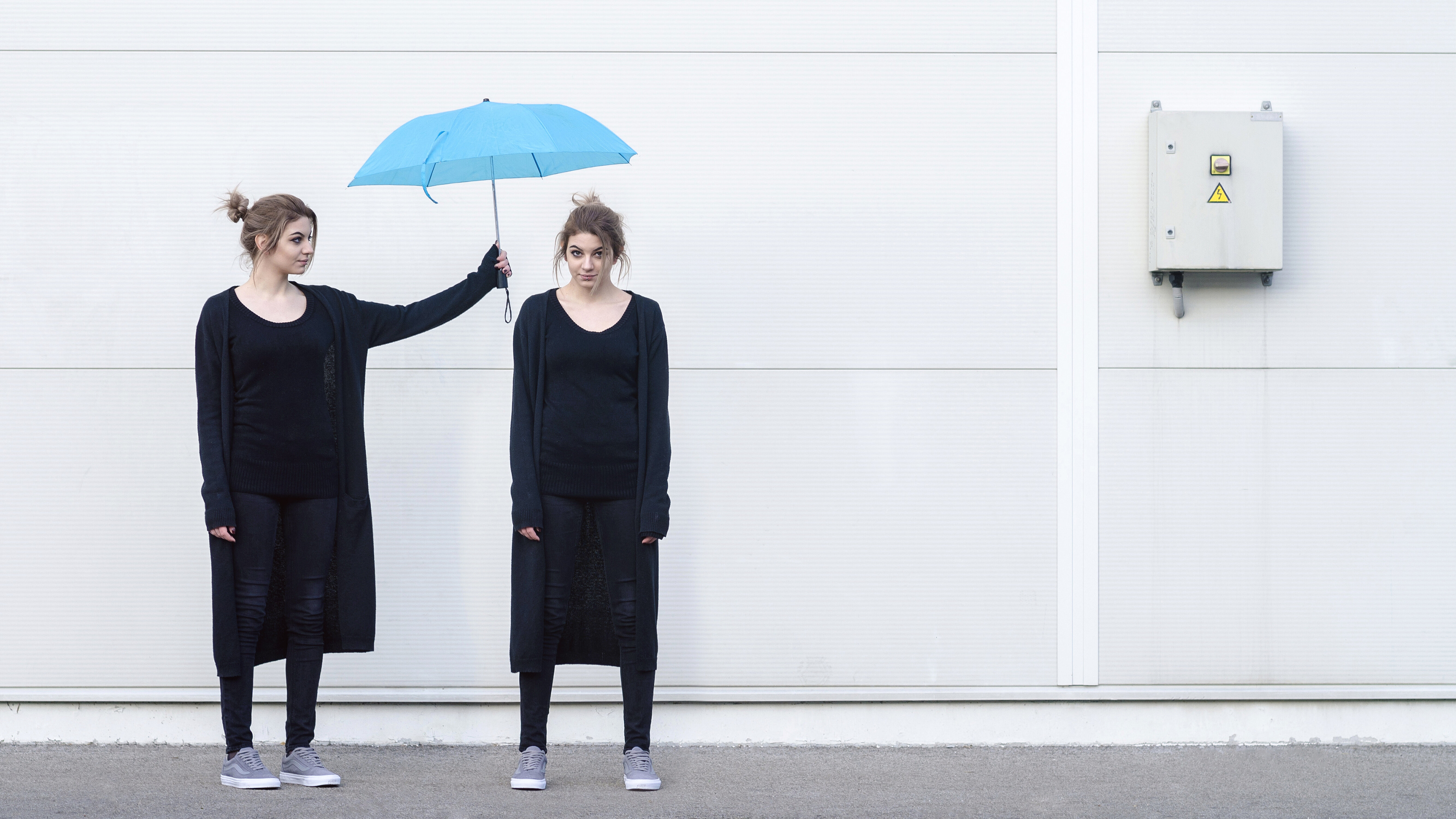 Eine junge Frau in schwarzer Kleidung hält einen blauen Regenschirm über den Kopf einer weiteren jungen Frau, die genauso aussieht wie die Frau, die den Regenschirm hält..