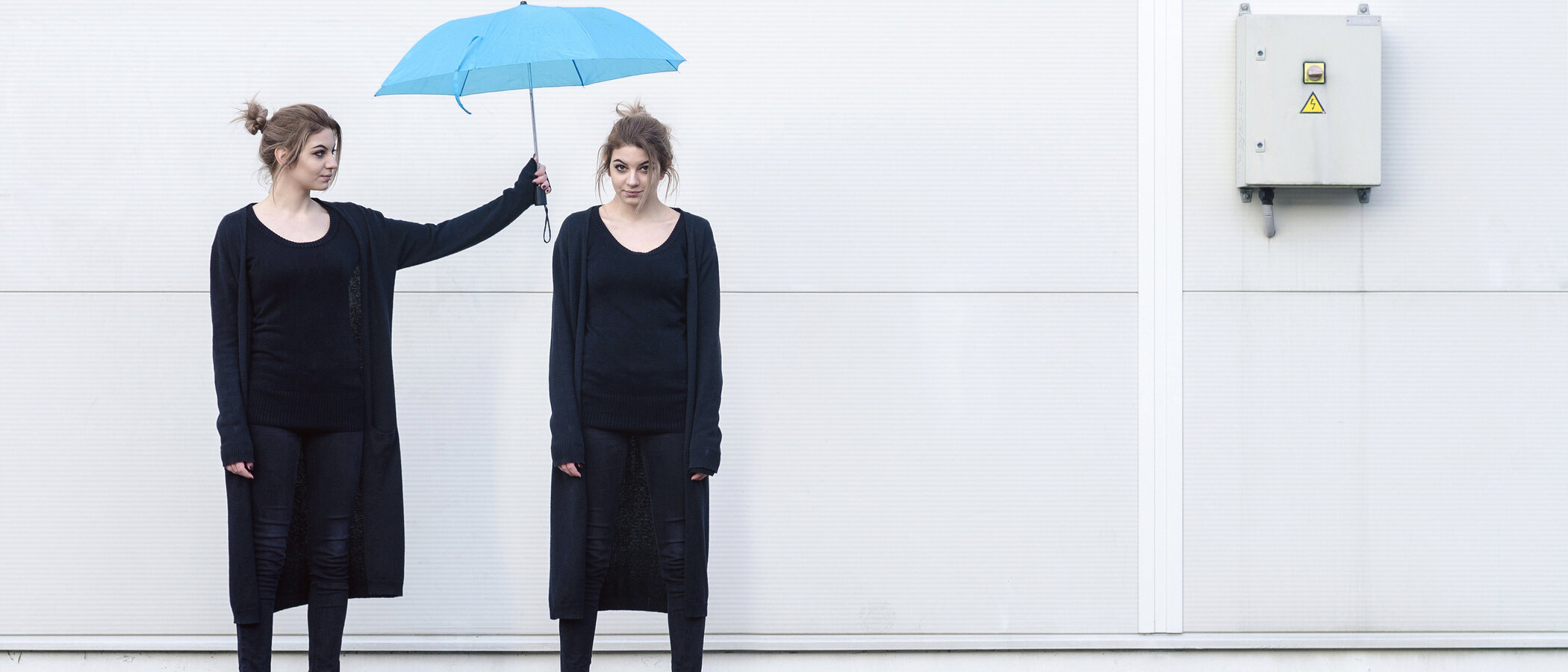 Eine junge Frau in schwarzer Kleidung hält einen blauen Regenschirm über den Kopf einer weiteren jungen Frau, die genauso aussieht wie die Frau, die den Regenschirm hält..