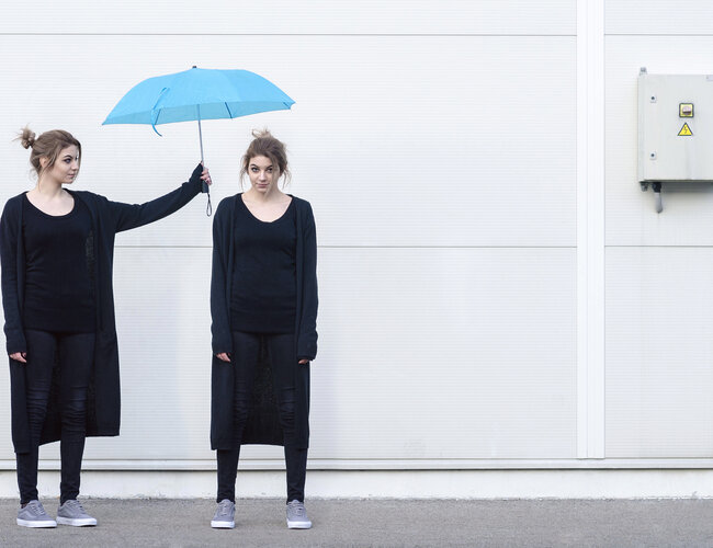 Eine junge Frau in schwarzer Kleidung hält einen blauen Regenschirm über den Kopf einer weiteren jungen Frau, die genauso aussieht wie die Frau, die den Regenschirm hält..