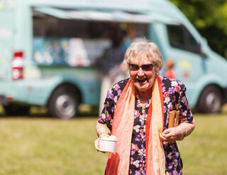 Seniorenbüro „Am Puls“ in Berlin: Agebote Eine lachende Seniorin läuft auf einer Wiese und hält in ihrer rechten Hand einen Eisbecher. Im Hintergrund ist ein Fahrzeug, aus dem heraus Eis verkauft wird.