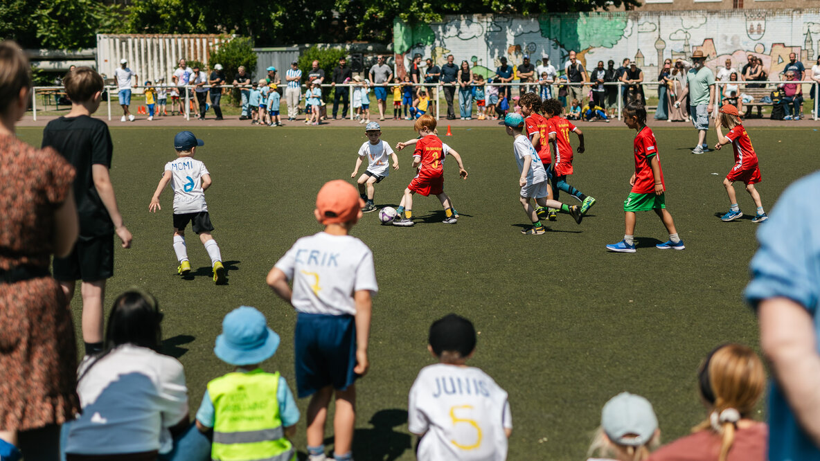 Auf einem Fußballfeld sind zwei Kinderteams zu sehen, die um den kämpfen. Im Vordergrund sitzen gespannt ein paar kleine Zuschauer*innen.