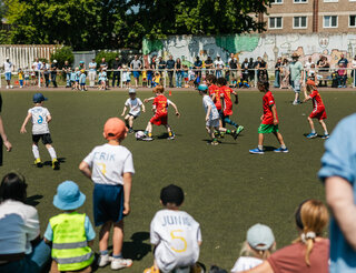 Familienfest und 10. Kicker-Fußballturnier der Humanistischen Kitas Auf einem Fußballfeld sind zwei Kinderteams zu sehen, die um den kämpfen. Im Vordergrund sitzen gespannt ein paar kleine Zuschauer*innen.