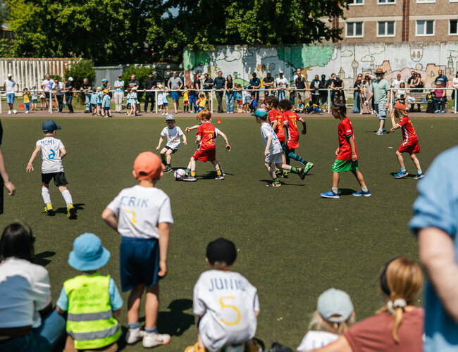 Auf einem Fußballfeld sind zwei Kinderteams zu sehen, die um den kämpfen. Im Vordergrund sitzen gespannt ein paar kleine Zuschauer*innen.