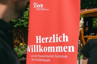 Humanistische Fachschule für Sozialpädagogik Eine Frau steht an einem Tresen. Ihre linke Hand ruht auf einem Ordner. Ihr gegenüber steht eine Person, von der nur die rechte Seite des Oberkörpers zu sehen ist. Ihre Hände ruhen auf dem Tresen. Im Hintergrund mittig ist ein rotes Roll-up, auf dem Folgendes zu lesen ist: „Herzlich willkommen an der Humanistischen Fachschule für Sozialpädagogik.“