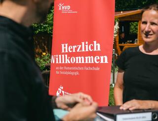 Humanistische Fachschule für Sozialpädagogik Eine Frau steht an einem Tresen. Ihre linke Hand ruht auf einem Ordner. Ihr gegenüber steht eine Person, von der nur die rechte Seite des Oberkörpers zu sehen ist. Ihre Hände ruhen auf dem Tresen. Im Hintergrund mittig ist ein rotes Roll-up, auf dem Folgendes zu lesen ist: „Herzlich willkommen an der Humanistischen Fachschule für Sozialpädagogik.“
