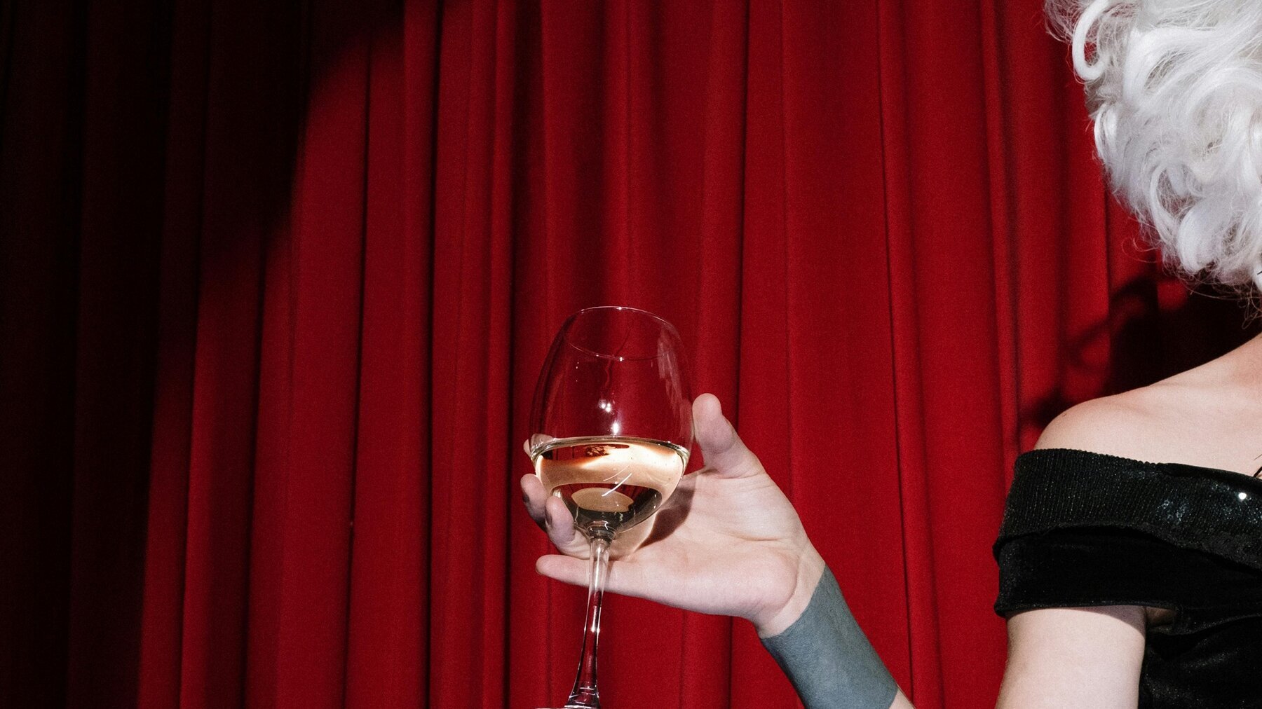 Eine Person in einem Kleid steht vor einem roten Vorhang und hält ein Weinglas in der Hand.