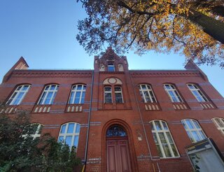 Alt-Buckow 17 Villa Zu sehen ist ein eine große Backsteinvilla von unten vor dem Eingang fotografiert. Dadurch ist der blaue Himmel zu sehen. Die Villa hat zwei Stockwerke, in der Mitte eine Art Turm und rechts und links jeweils sechs große Fenster.