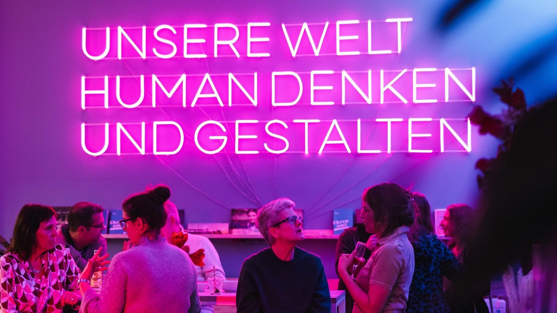 Im Haus des HUMANISMUS in der Potsdamer Straße in Berlin sind Leuchtbuchstaben an der Wand zu sehen. Zu lesen ist in Großbuchstaben: „Unsere Welt human denken und gestalten“. Davor sitzen Menschen an einem Tresen und unterhalten sich.