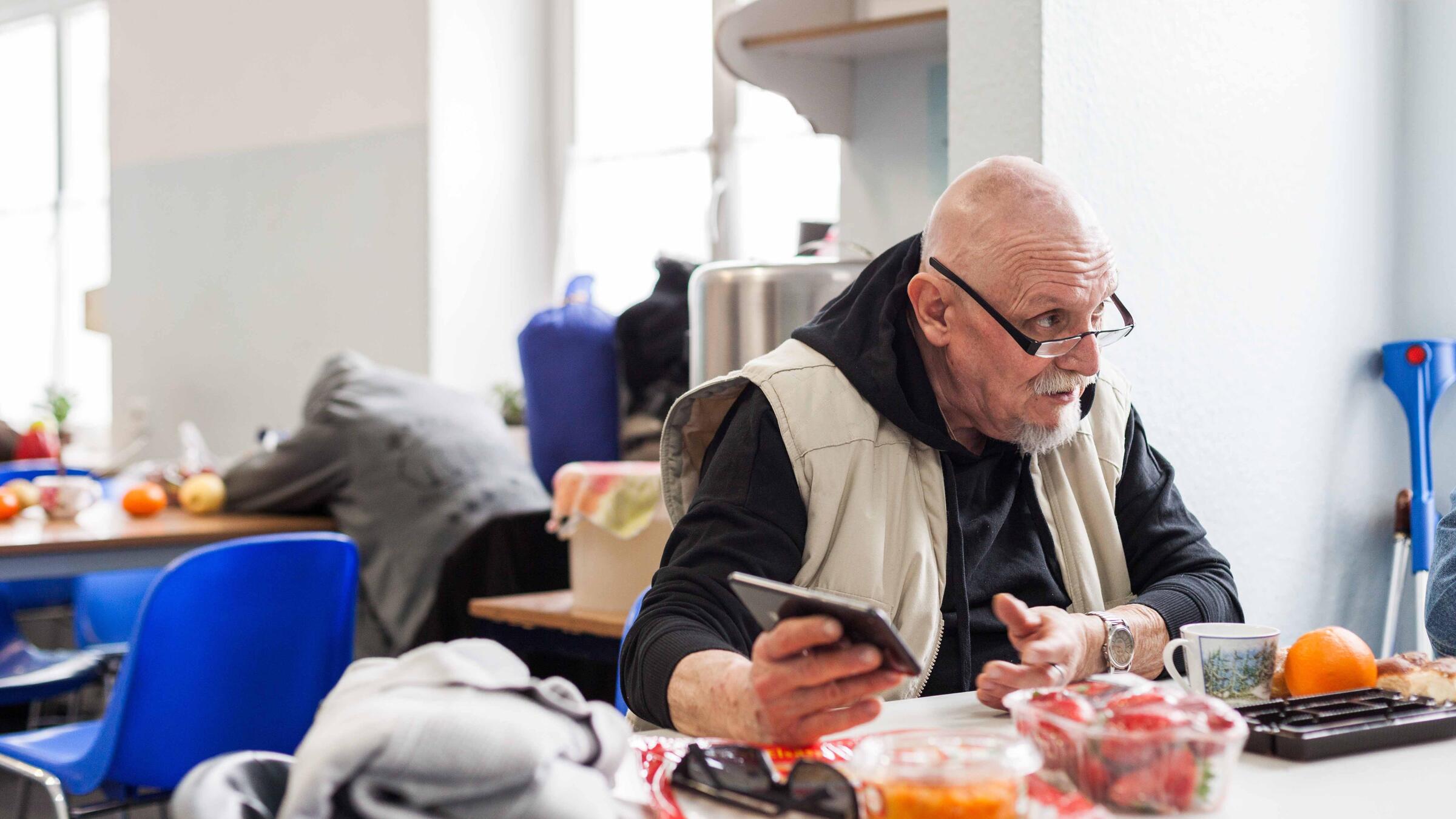 Ein Mann in einer beigen Weste und mit einer Brille sitzt an einem Tisch. Vor ihm steht Essen. In der rechten Hand hält er ein Handy und er scheint mit jemanden zu reden. Im Hintergrund sieht man einen weiteren Tisch und verschwommen eine Person, die daran sitzt.