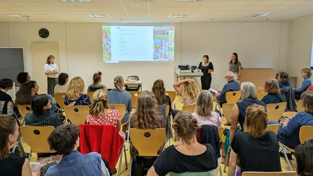 Personen sind von hinten sitzend in einem Raum auf Stühlen zu sehen. Sie hören drei Dozierenden zu, die im Hintergrund stehen. Ein Beamer projiziert ein Bild oben mittig auf die Wand, auf die die Personen blicken.