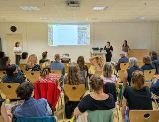 Humanistische Lebenskunde: Rückblick 1. Barcamp für Lebenskunde-Lehrkräfte 2025 Personen sind von hinten sitzend in einem Raum auf Stühlen zu sehen. Sie hören drei Dozierenden zu, die im Hintergrund stehen. Ein Beamer projiziert ein Bild oben mittig auf die Wand, auf die die Personen blicken.