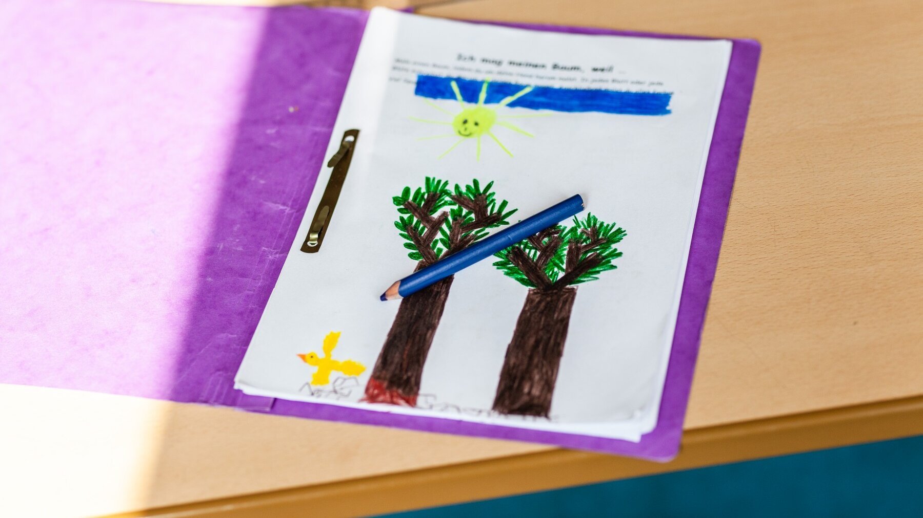 Auf einer Tischplatte liegt ein geöffneter lila Papphefter. Darin ist das obere Arbeitsblatt zu sehen. Darauf wurden ein Himmel, eine Sonne, zwei Bäume und links unten neben dem Baum links ein Vogel mit Filzstiften gemalt. Auf dem Blatt liegt quer ein dunkelblauer Buntstift.