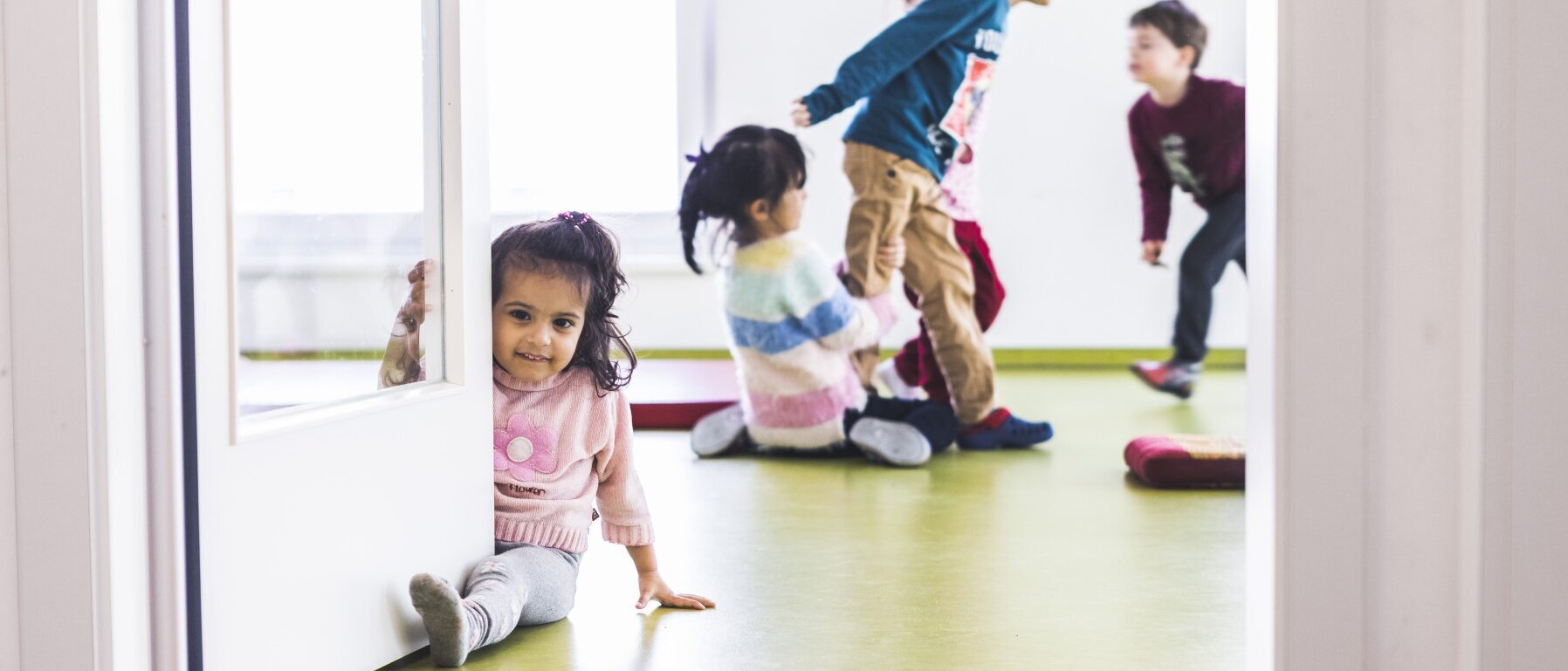 In einem Raum mit lindgrünem Fußboden, weißen Wänden und weißer Glastür spielen fünf Kinder.
