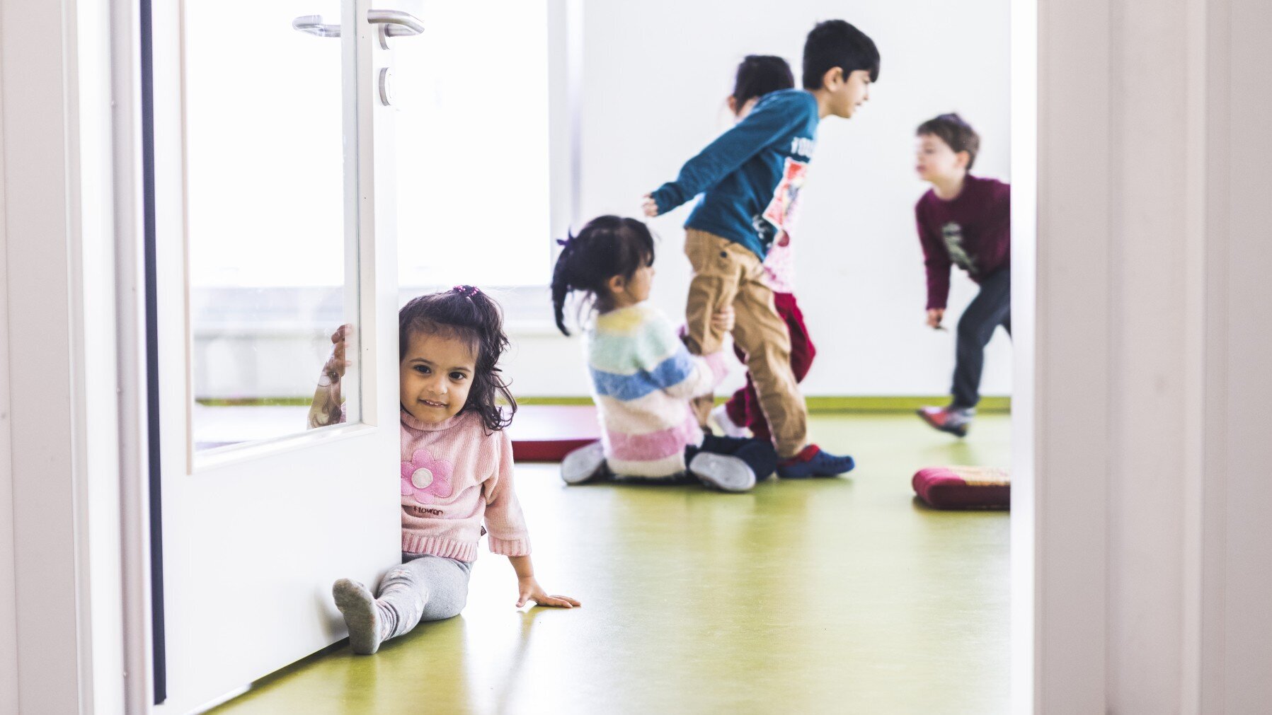 In einem Raum mit lindgrünem Fußboden, weißen Wänden und weißer Glastür spielen fünf Kinder.