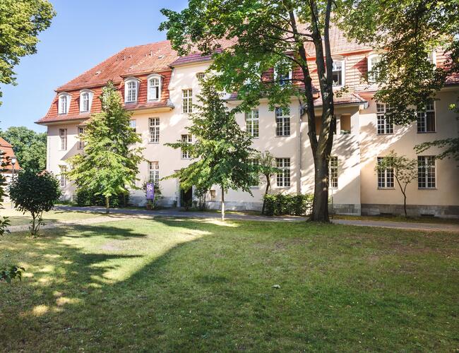 Zu sehen ist eine große grüne Wiese mit Bäumen. Hinten steht das Hospiz LudwigPark: ein großes helles und breites Haus mit vielen Fenstern.