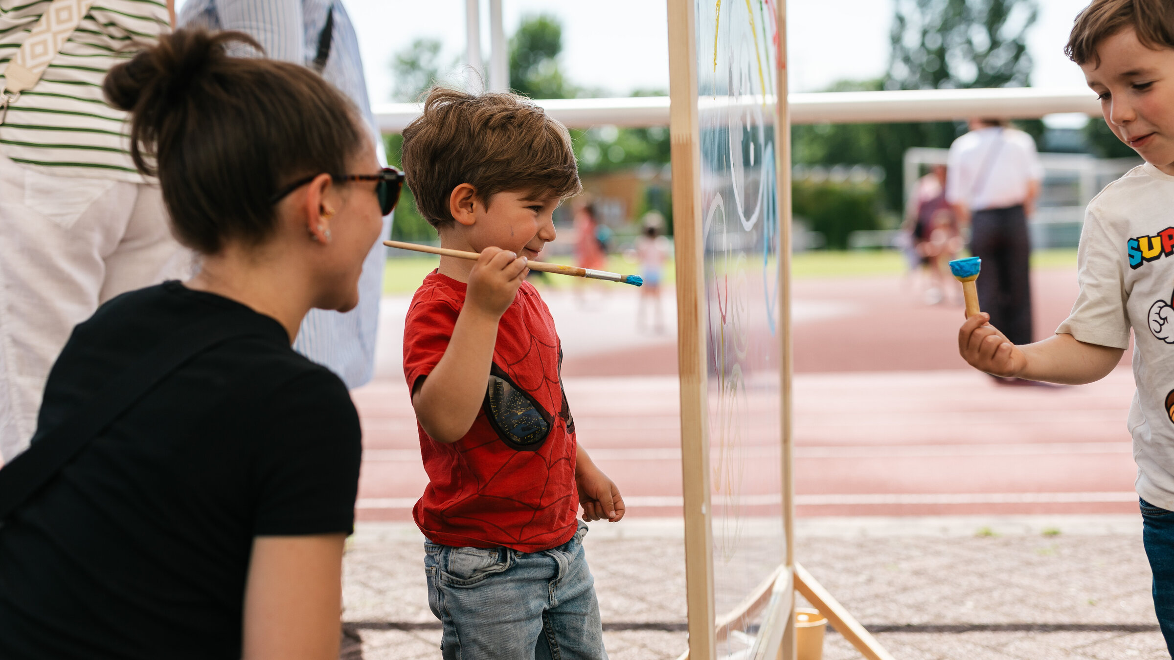 Ein kleiner Junge malt etwas auf einer transparenten Oberfläche, die vor ihm aufgespannt ist. Neben ihm hockt eine Frau mit Sonnenbrille.