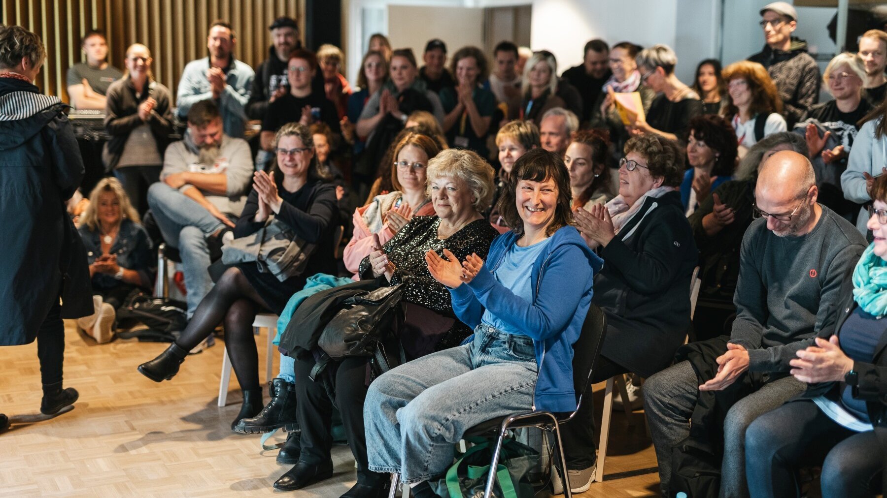 In mehreren Reihen sitzende sowie dahinter stehende Personen klatschen.