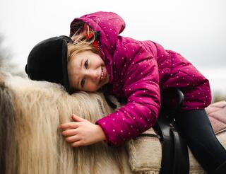 Kinderhospiz Berliner Herz: Begegnungen mit Tieren Ein Mädchen mit Reiterhelm und Reitstiefeln sitzt beziehungsweise liegt bäuchlings auf dem Rücken eines Ponys im Sattel und lächelt.