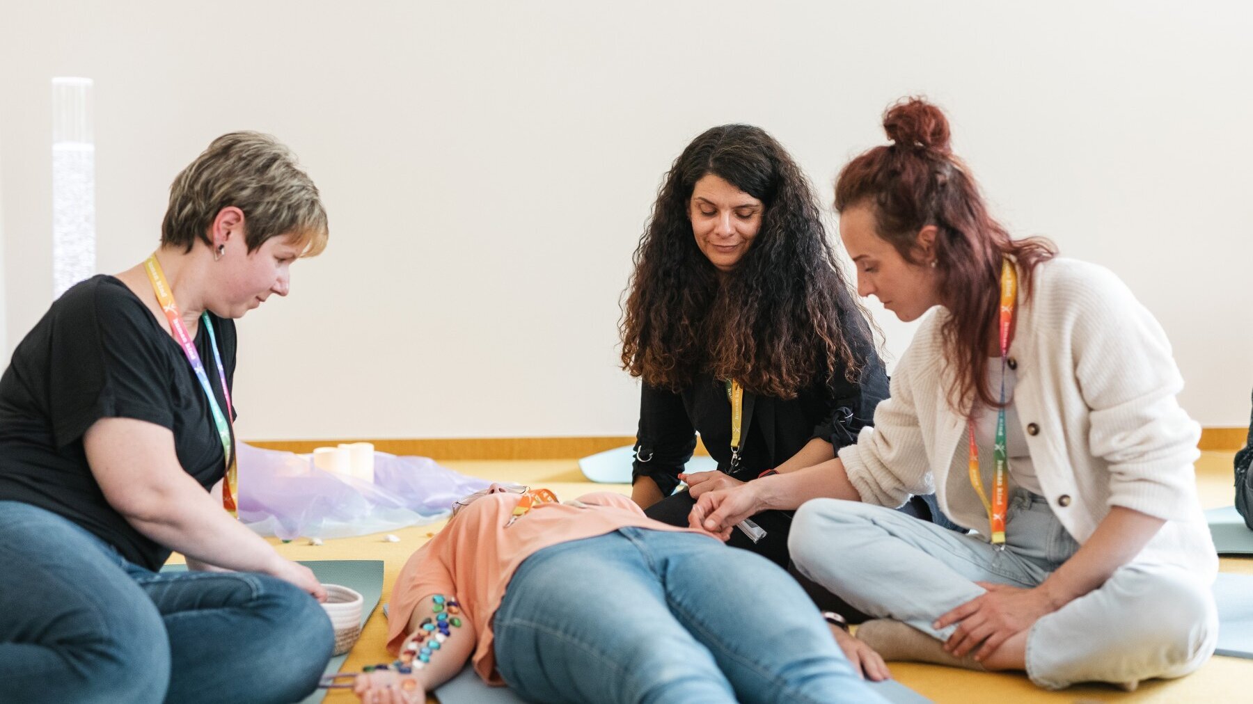 Eine Frau liegt auf einer Matte. Links von ihr sitzt eine Frau, rechts von ihr sitzen zwei Frauen. Auf den Unterarmseiten innen der liegenden Frau werden von den anderen Steinchen platziert.