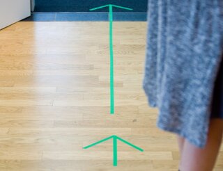 Über die Kontakt- und Informationsstelle für Selbsthilfe in Berlin-Pankow Auf einem Parketboden sind Pfeile aus grünem Klebeband. Rechts sind Beine einer Person zu sehen.