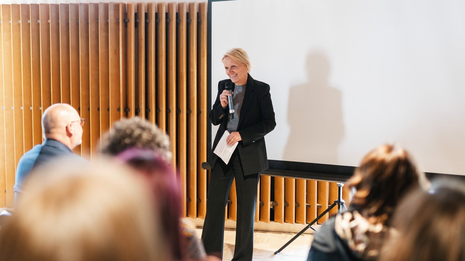 Katrin Raczynski, Vorstandsvorsitzende des Humanistischen Verbandes Berlin-Brandenburg, spricht in ein Mikrofon. Im Vordergrund sind sechs Hinterköpfe sitzender Personen zu sehen.
