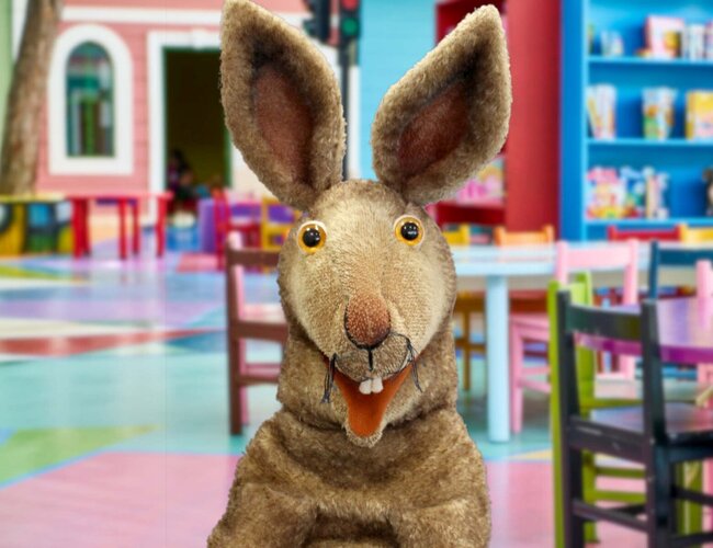 Eine Hasenhandpuppe ist im Vordergrund zu sehen. Im Hintergrund ist alles bunt: der Fußboden, die Kindertische und Kinderstülel, ein Regal mit Büchern.