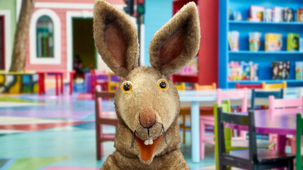 Eine Hasenhandpuppe ist im Vordergrund zu sehen. Im Hintergrund ist alles bunt: der Fußboden, die Kindertische und Kinderstülel, ein Regal mit Büchern.