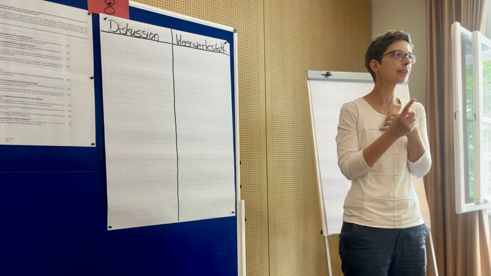 Zu sehenist eine Frau die rechts neben einem Board steht und etwas erzählt. Sie hat kurze Haare, eine Brille und ein weißes Oberteil. Auf dem Board ist eine Tabelle unterteilt in: Diskussion und Ideenwerkstatt.