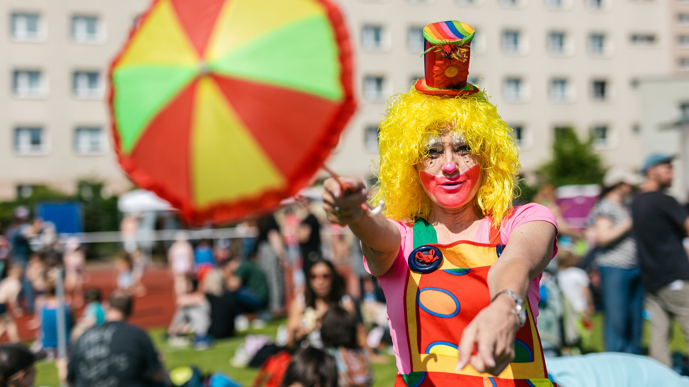 Ein buntgekleideter Clown mit gelber Perücke häkt einen bunten Schirm vor sich und schaut in die Kamera.