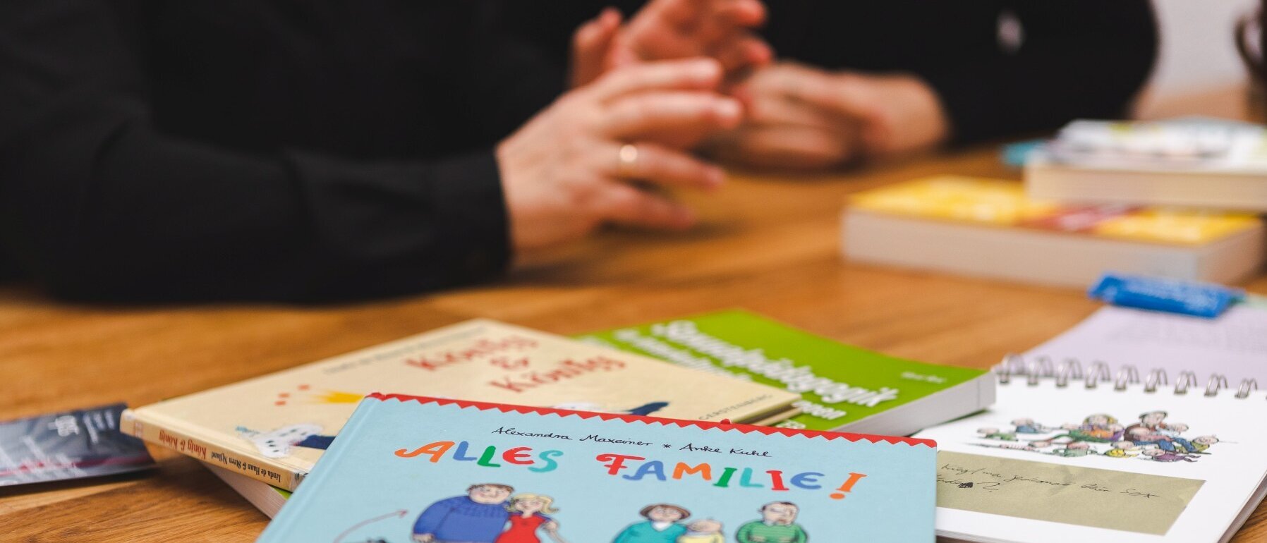 Auf einem Tisch liegen Bücher, auf dem vorn steht: „Alles Familie!“ Im Hintergrund sind von zwei Frauen die Hände, Unterame und der Oberkörper bis zum Hals zu sehen.