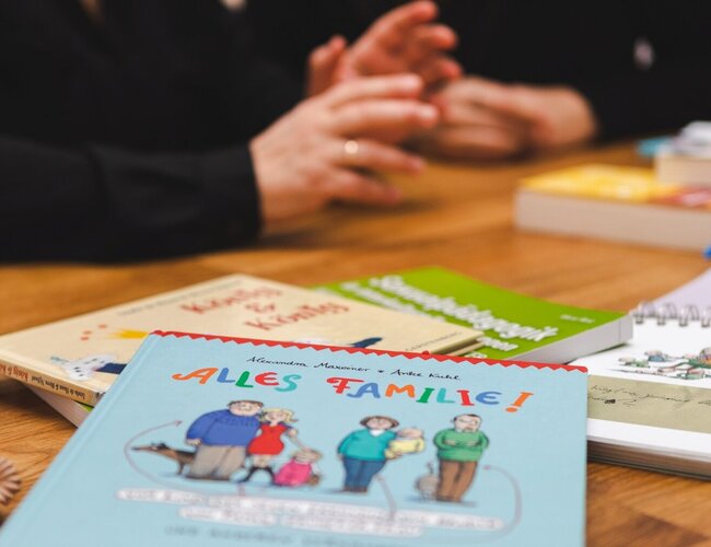 Auf einem Tisch liegen Bücher, auf dem vorn steht: „Alles Familie!“ Im Hintergrund sind von zwei Frauen die Hände, Unterame und der Oberkörper bis zum Hals zu sehen.