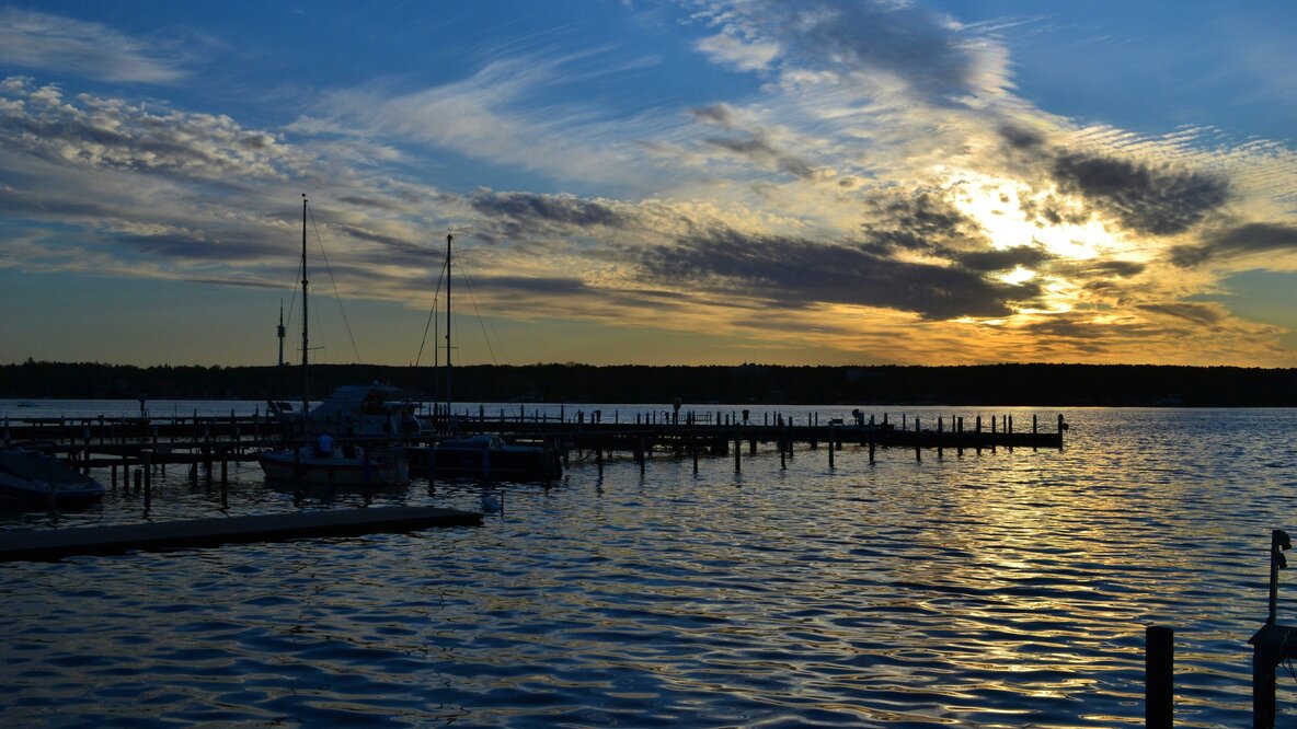 Auf den Bild sieht man den Wannsee bei Sonnenuntergang. Von links ragt ein langer Steg in den See rein, an dem ein weißes Bood angelegt ist. Der Sonnenuntergang spiegelt sich auf der Wasseroberfläche.