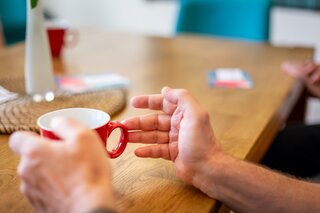 KIS: Kontakt- und Informationsstelle für Selbsthilfe in Berlin-Pankow Eine linke Hand hält eine auf einem Tisch stehende Tasse, die rechte ist horizontal auf der Tischplatte.