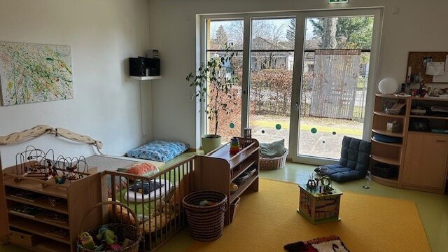 In einem Raum mit Teppich und einer Gitterwand, die den Raum teilt, befinden sich Schränke und Körbe mit Spielzeug sowie Kissen.