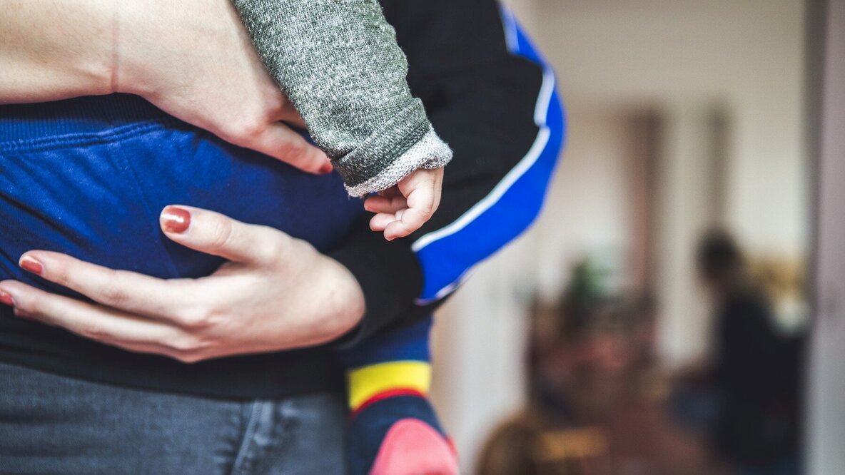 Eine Erzieherin trägt ein Kleinkind, nur die Mitte der Körper ist zu sehen. Rechts im Hintergrund sind verschwommen weitere Personen zu sehen.