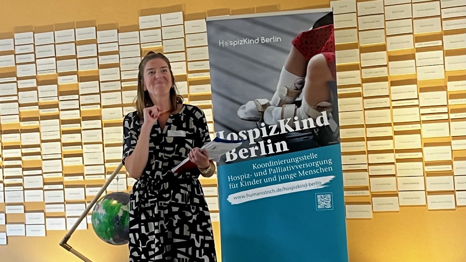 Vor einer gelben Wand steht eine Frau neben einem RollUps von HospizKind Berlin. Sie hält ein Heft in der Hand und schaut lächelnd nach vorne.