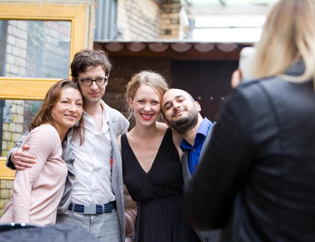 Vier Menschen in festlicher Kleidung stehen nebeneinander im Freien und werden von einer Frau mit dem Handy fotografiert.