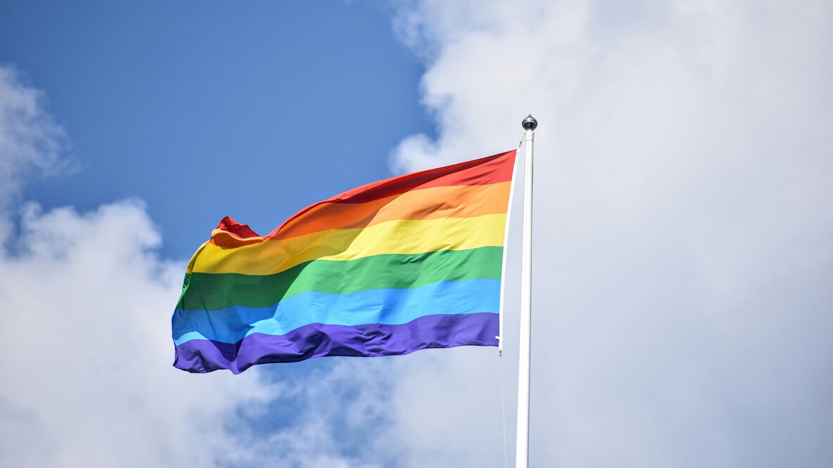 Eine Regenbogenfahne weht an einem Fahnenmast vor einem blauen Himmel mit weißen Wolken.