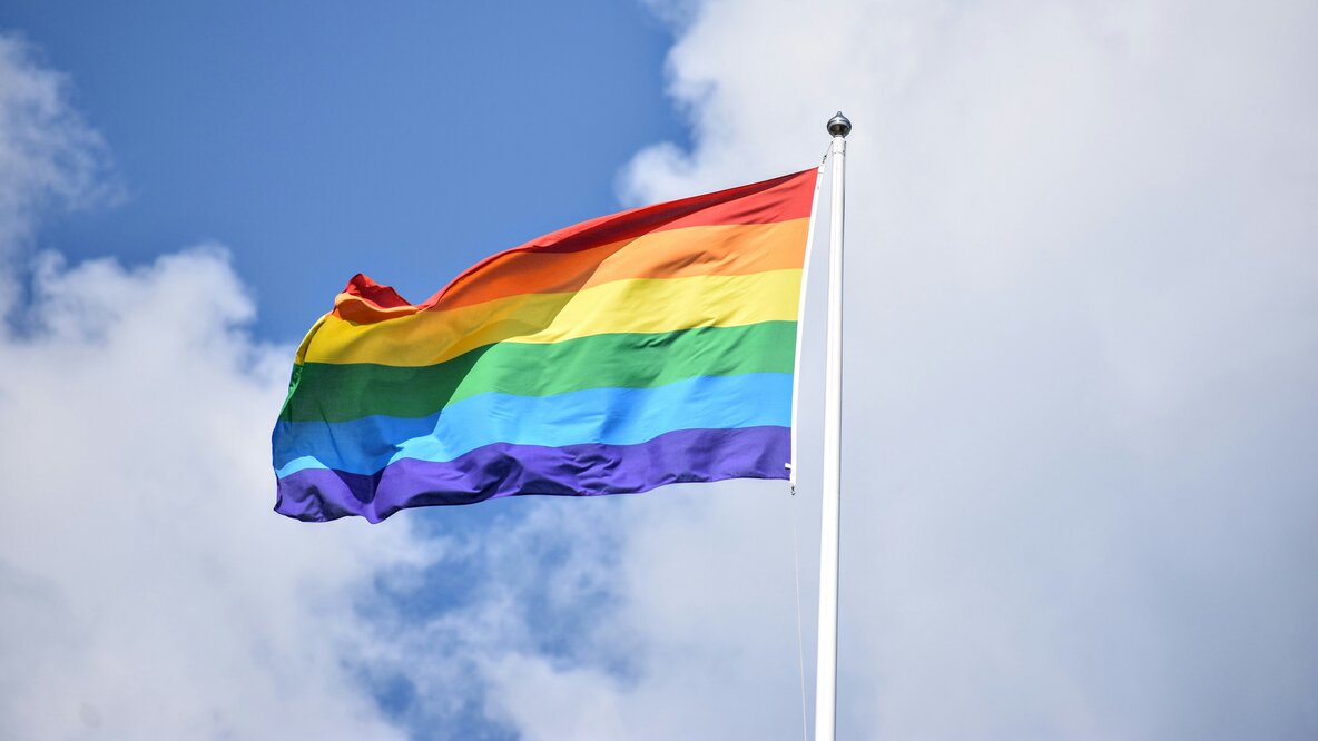 Eine Regenbogenfahne weht an einem Fahnenmast vor einem blauen Himmel mit weißen Wolken.