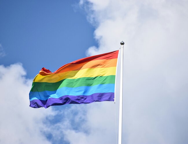 Eine Regenbogenfahne weht an einem Fahnenmast vor einem blauen Himmel mit weißen Wolken.
