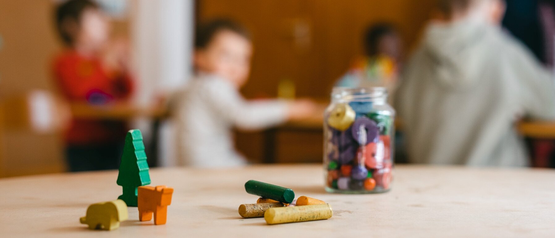 Auf einem Tisch stehen lHolzfiguren und ein Glas mit Fettstiften, einige Stifte liegen vor dem Glas. Im Hintergrund sind unscharf fünf Kinder zu sehen.
