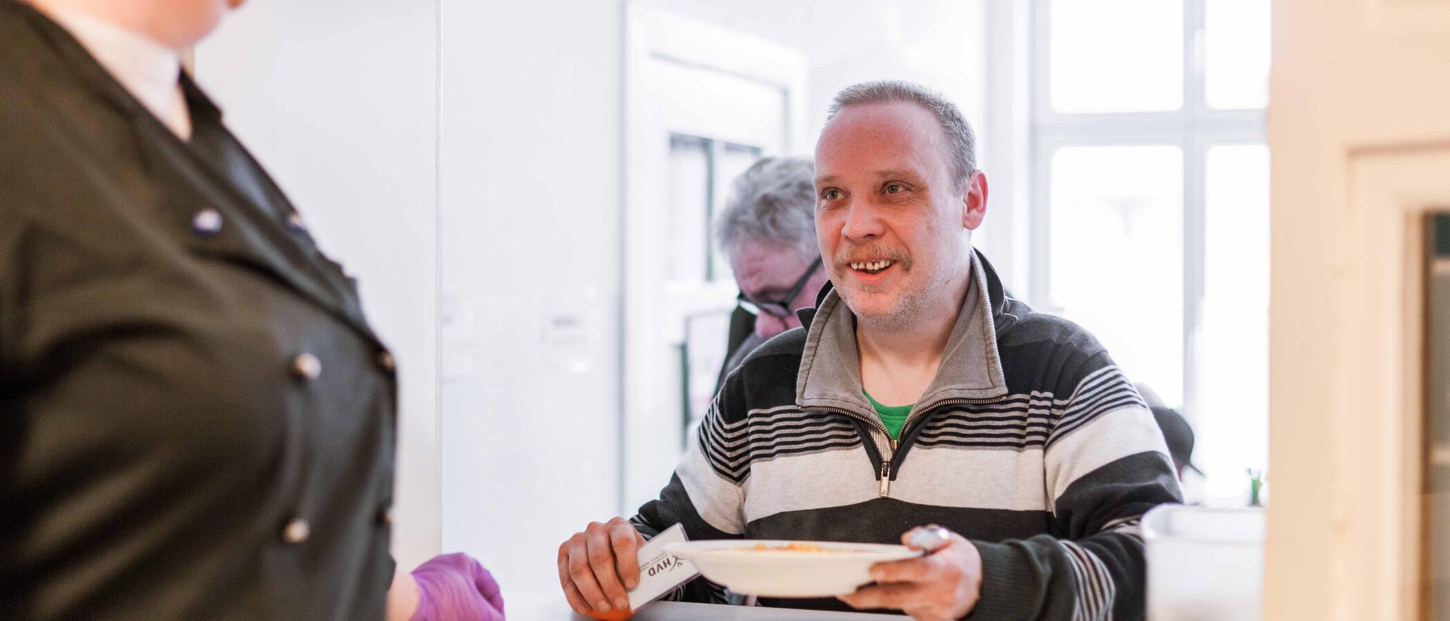 Links steht angereissen eine Frau in einer schwarzen Kochjacke und schaut druch die Luke der Essensausgabe auf einem Mann, der sich gerade einen Teller mit warmen Essen und eine Mandarine von der Theke nimmt. Sie lächeln sich an. 