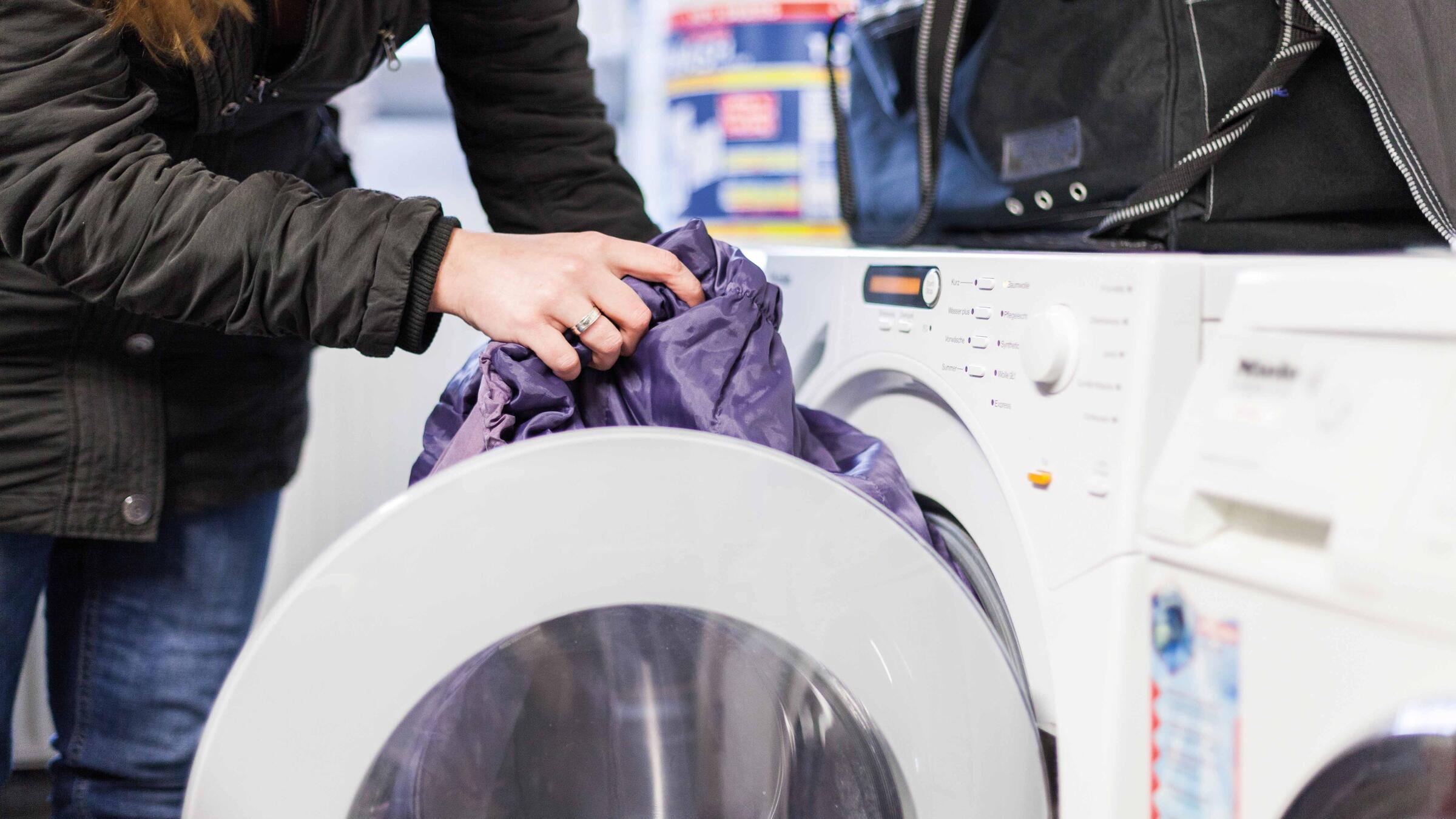 Zu sehen ist eine Person in einer schwarzen Jacke, die Kleidung in eine Waschmaschine steckt. Der Kopf der Person ist nicht zu sehen.