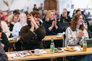 Humanistischer Verband Berlin-Brandenburg: Mitglieder und Freund*innen Menschen sitzen an Tischen in Reihen in einem Raum. In der Mitte ist ein Mann zu sehen, der klatscht und herzlich lacht.