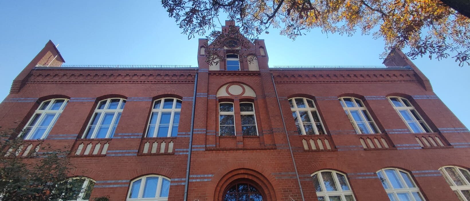 Zu sehen ist ein eine große Backsteinvilla von unten vor dem Eingang fotografiert. Dadurch ist der blaue Himmel zu sehen. Die Villa hat zwei Stockwerke, in der Mitte eine Art Turm und rechts und links jeweils sechs große Fenster.