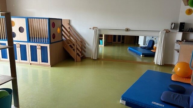 In einem Raum befinden sich Matten, Sitzkissen, Bälle und Klettermöglichkeiten. Hinten rechts unten an der Wand ist ein Spiegel befestigt, der sich mit Vorhängen verbergen lässt.